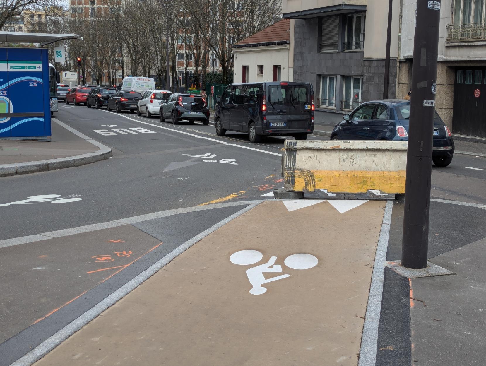 Dans un virage en fin de couloir de bus, une piste cyclable est visible dans la continuité du couloir, un bloc de chantier en béton se trouve entre le couloirs de bus et la piste cyclable.