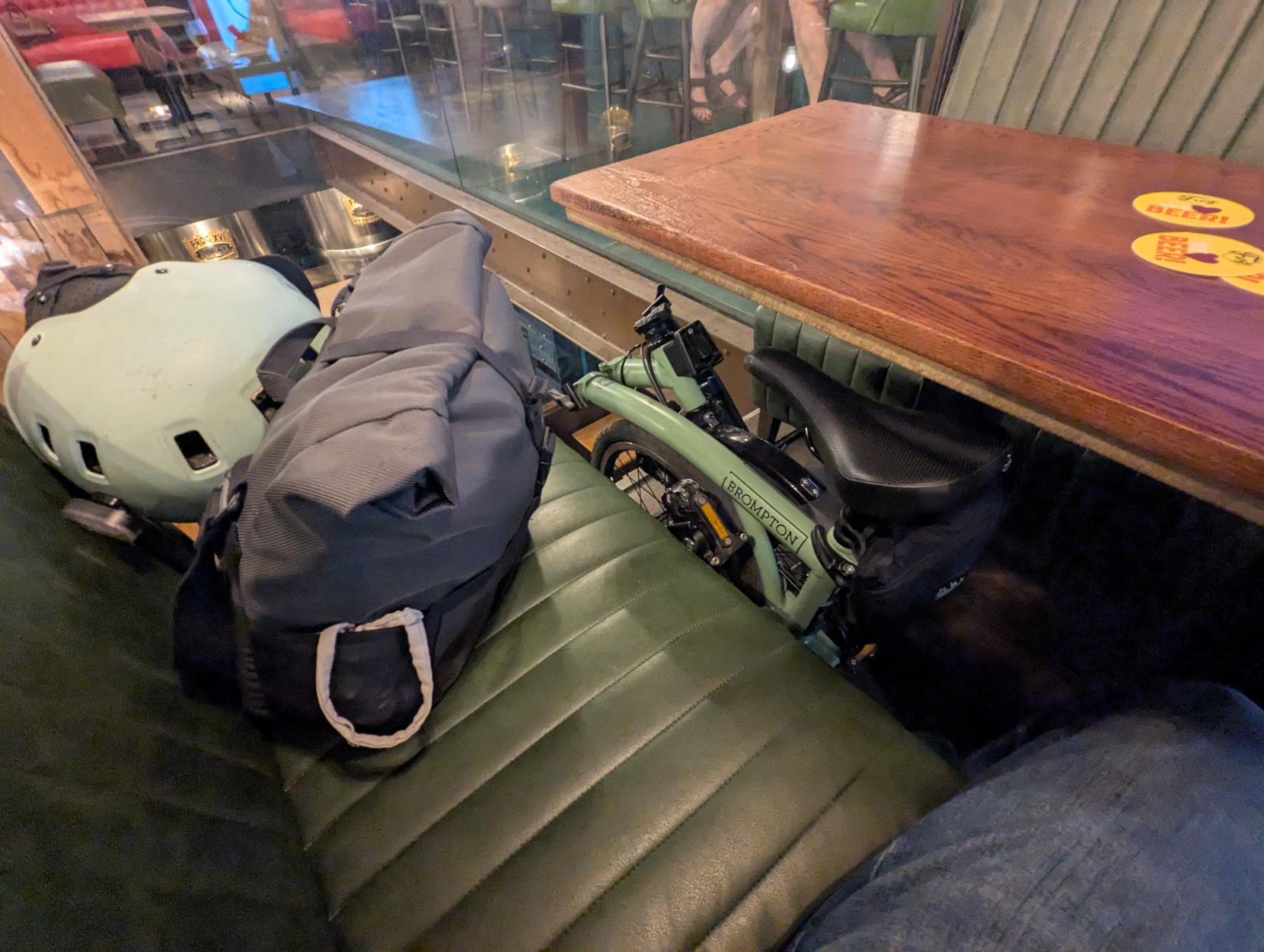 Un Brompton plié, calé entre une table de bar et une banquette. Sur la banquette se trouve une sacoche et un casque.