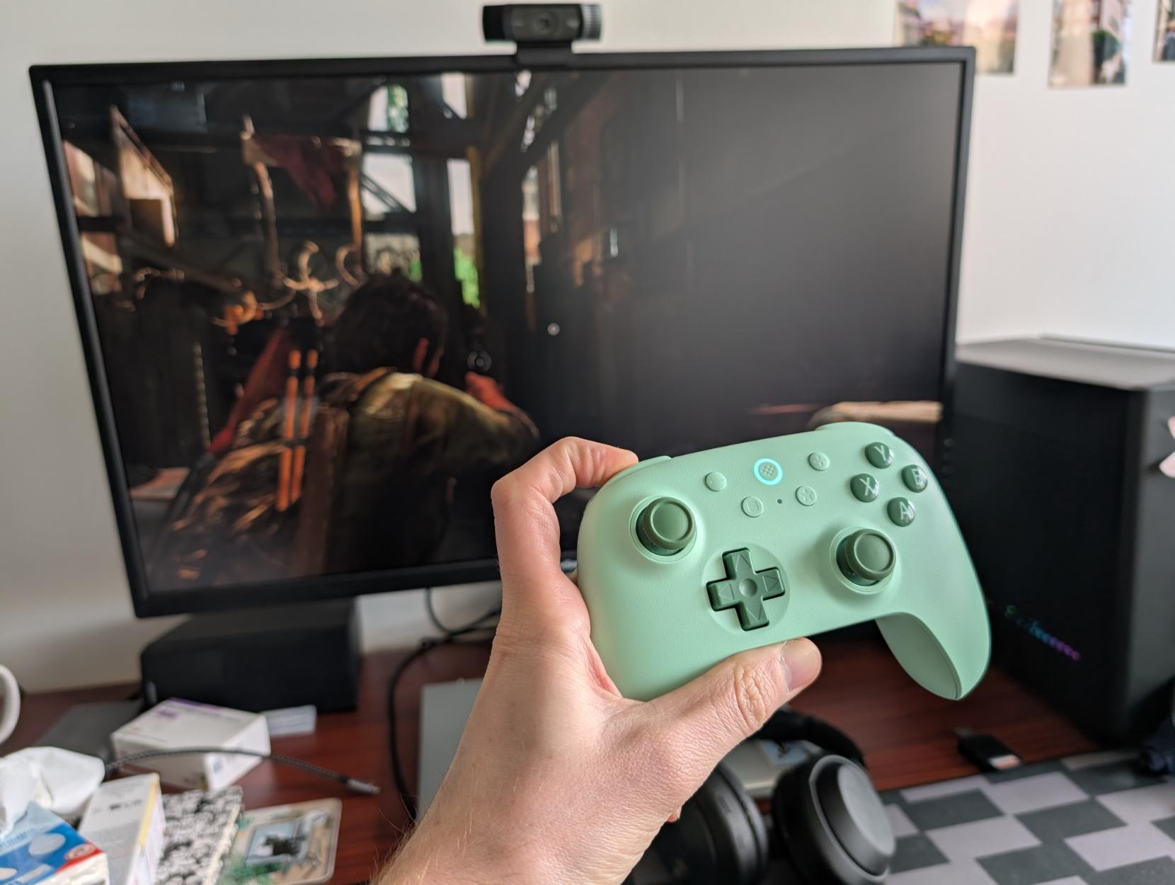 A green game controller in front of a PC and its monitor. The monitor shows a frame of the game The Last of Us.